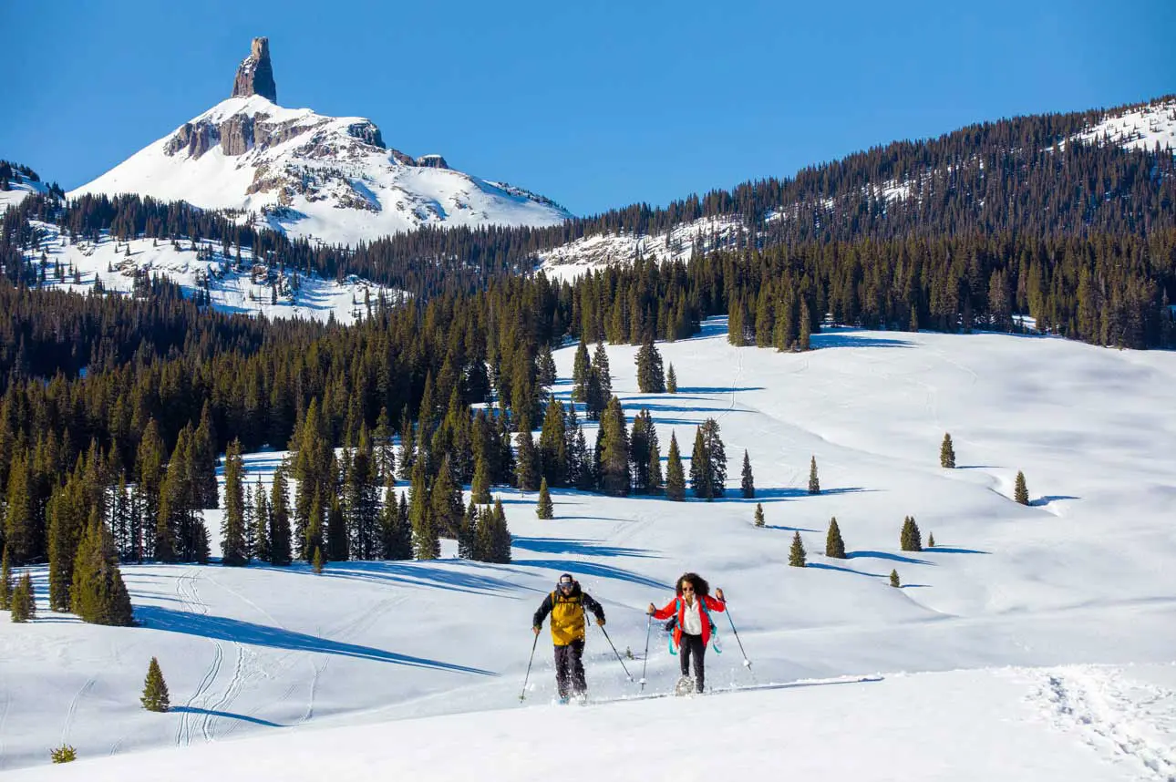 Two people with snowshoes and poles cross a snow field with a snow-covered peak in the distance under blue skies